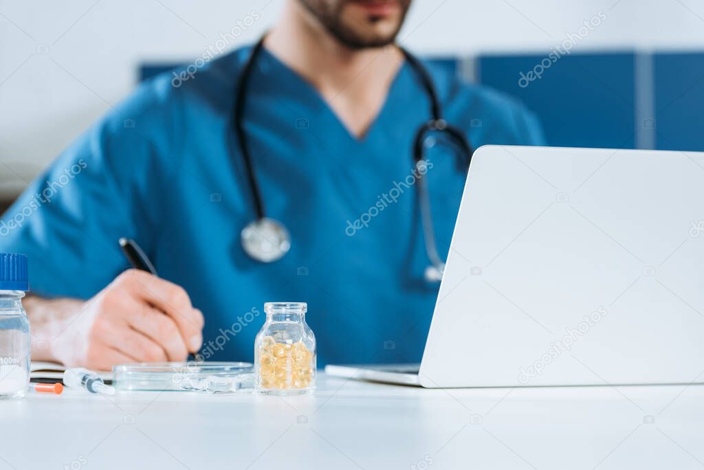 selective focus of young doctor writing near laptop and container with medicines