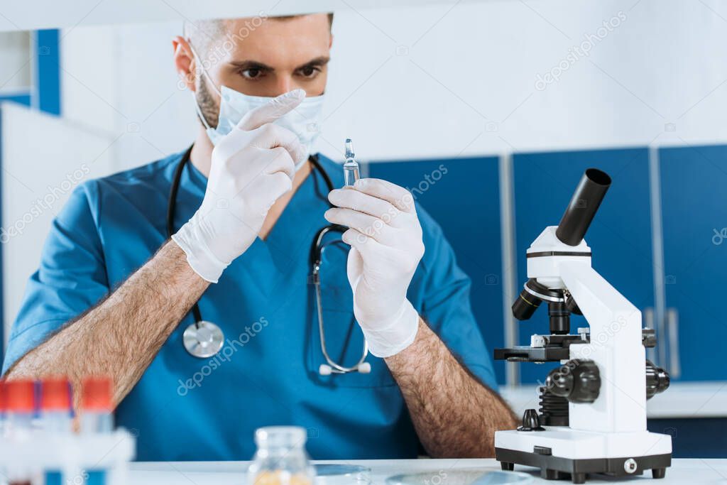 selective focus of biologist in medical mask and latex gloves looking at ampule with medicine near microscope