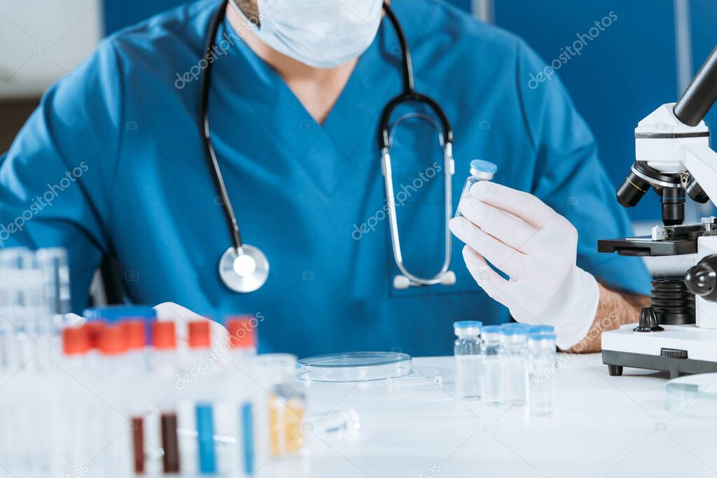 cropped view of biologist in latex gloves holding glass container with vaccine near test tubes