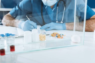 cropped view of veterinarian in medical mask and lates gloves writing near containers with medicines and white mouse in glass box clipart