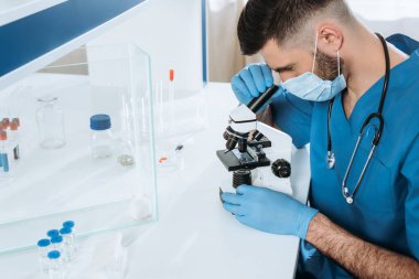Tıbbi maskeli genç bir biyolog ve cam kutuda mikroskopla analiz yapan eldivenler ve içinde ilaç bulunan kaplar olan beyaz bir fare.