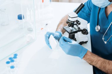 Tıbbi maskeli bir biyolog ve test kutusunu tutan eldivenleri incelerken cam kutunun içindeki beyaz farenin yanında mikroskop kullanarak analiz yapıyor.