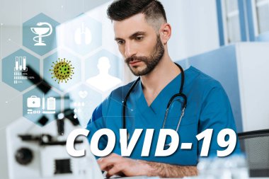 Mikroskop, koronavirüs illüstrasyonu yakınlarındaki dizüstü bilgisayarda yazan konsantre doktorun seçici odak noktası