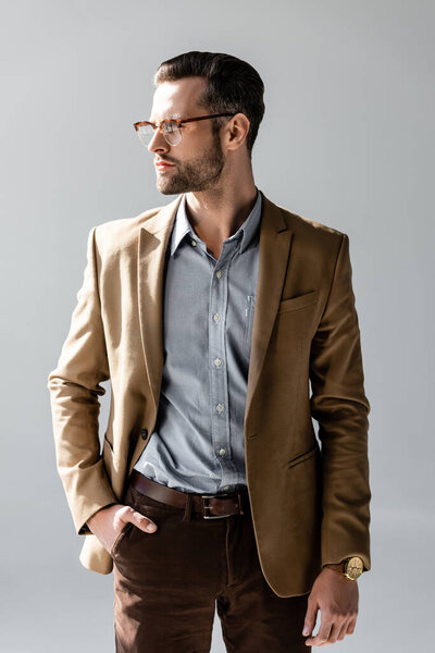 handsome businessman in glasses looking away and standing with hand in pocket isolated on grey 