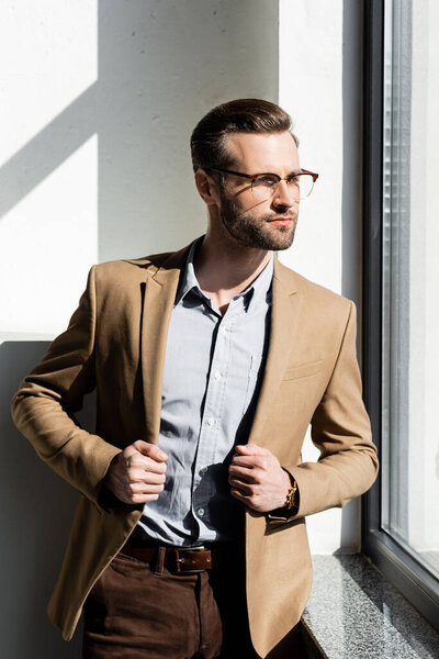 bearded businessman looking at window and touching blazer 