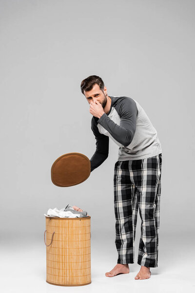 bearded man touching nose while standing near smelly dirty laundry on grey 