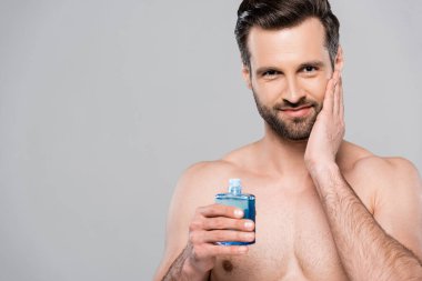 Tıraş losyonundan sonra mavi şişeyi tutan mutlu adam ve gri renkte izole edilmiş yüzlere dokunan adam.  