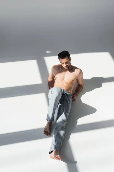 穿着蓝色牛仔裤坐在灰色上的肌肉发达的男人身上的阳光 — 图库照片