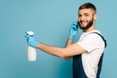 side view of happy cleaner in uniform and rubber gloves holding spray detergent and talking on smartphone on blue background