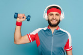 šťastný stylový sportovec ve sluchátkách cvičení s činka na modrém pozadí