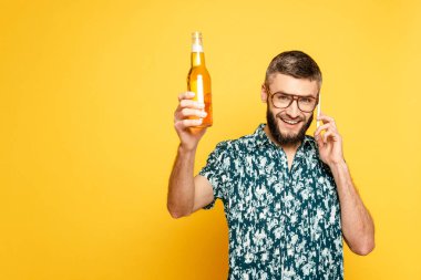 Gözlüklü mutlu sakallı adam ferahlatıcı bir bira şişesiyle sarıda akıllı telefondan konuşuyor.