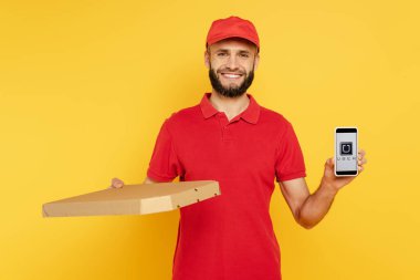 KYIV, UKRAINE - 30 Mart 2020: kırmızı üniformalı gülümseyen sakallı teslimatçı pizza kutulu akıllı telefonu sarı üzerine uygulamalı gösteriyor.