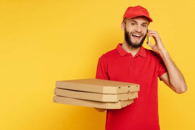 Kırmızı üniformalı gülümseyen sakallı teslimatçı elinde pizza kutularıyla sarı telefonda konuşuyor.