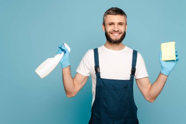 Mavi arka planda deterjan ve sünger tutan üniformalı mutlu bir temizlikçi ve lastik eldiven.