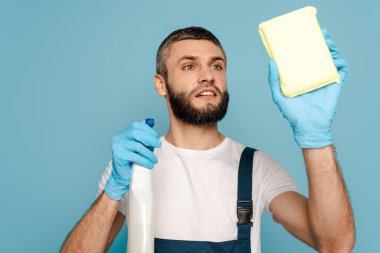 Mavi arka planda deterjan ve sünger kullanan üniformalı ve lastik eldivenli temizleyici.
