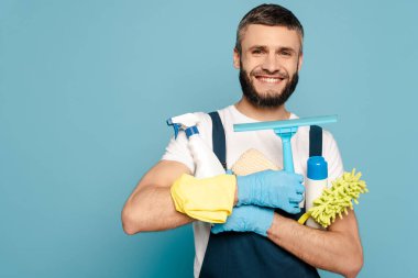 Üniformalı mutlu bir temizlikçi ve mavi arka planda temizlik malzemeleri tutan lastik eldivenler.