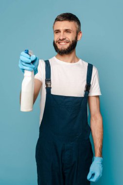 Üniformalı mutlu bir temizlikçi ve mavi arka planda sprey deterjan tutan lastik eldivenler.
