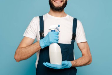 Üniformalı mutlu temizlikçi manzarası ve mavi arka planda sprey deterjan tutan lastik eldivenler.