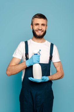 Üniformalı mutlu bir temizlikçi ve mavi arka planda sprey deterjan tutan lastik eldivenler.