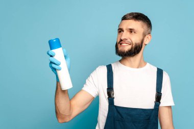 Üniformalı mutlu temizlikçi ve mavi arka planda oda spreyi tutan lastik eldiven.