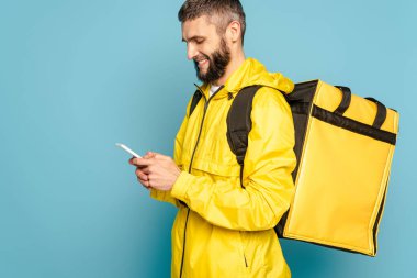 Sırt çantalı sarı üniformalı gülümseyen teslimatçı mavi arka planda akıllı telefon kullanıyor.