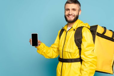 Sırt çantalı mutlu teslimatçı mavi arka planda boş ekranlı akıllı telefon gösteriyor.