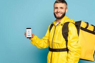 KYIV, UKRAINE - MARCH 30, 2020: happy deliveryman in yellow uniform with backpack showing smartphone with messenger app on blue background clipart