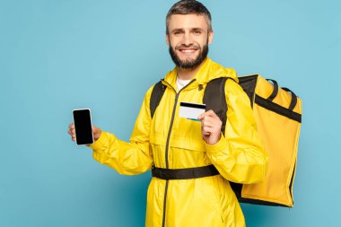 Sarı üniformalı mutlu teslimatçı mavi arka planda kredi kartı tutarken elinde boş ekranlı akıllı telefonu gösteriyor.