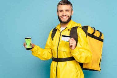 Sarı üniformalı mutlu teslimatçı, mavi arka planda kredi kartı tutarken moda alışveriş uygulamalı akıllı telefonu gösteriyor.