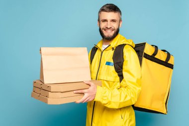 smiling deliveryman in yellow uniform with backpack holding paper package and pizza boxes on blue background clipart