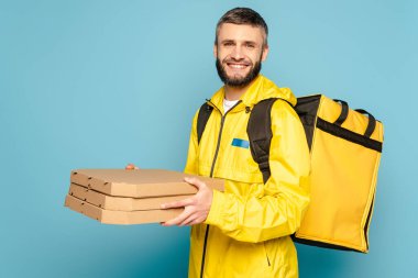 Sırt çantalı sarı üniformalı gülümseyen teslimatçı mavi arka planda pizza kutuları tutuyor.