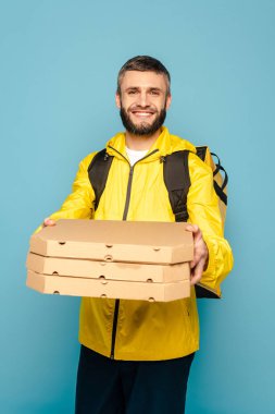 smiling deliveryman in yellow uniform with backpack holding pizza boxes on blue background clipart