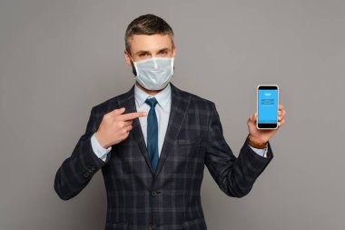 KYIV, UKRAINE - 30 Mart 2020: Sağlık maskeli yakışıklı işadamı gri arka planda Skype uygulamalı akıllı telefonu işaret ediyor