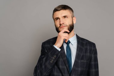 Takım elbiseli yakışıklı sakallı iş adamı gri arka planda çeneye dokunuyor.