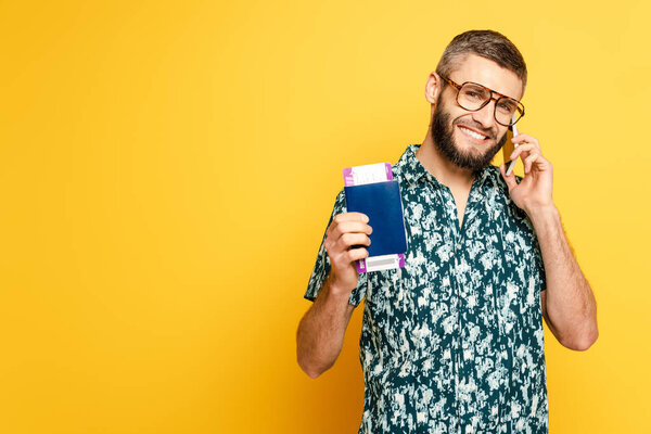 smiling bearded guy in glasses with passport and ticket talking on smartphone on yellow