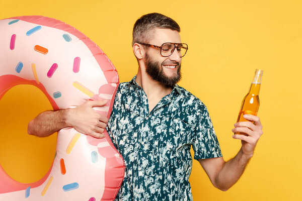 Улыбающийся бородатый парень в очках с кольцом из пончиков и пивом на желтом
