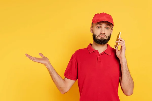 黄色のスマートフォンで話す赤い制服を着た混乱した髭の配達人 — ストック写真