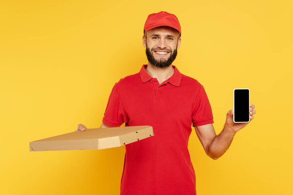 smiling bearded delivery man in red uniform with pizza box and smartphone on yellow