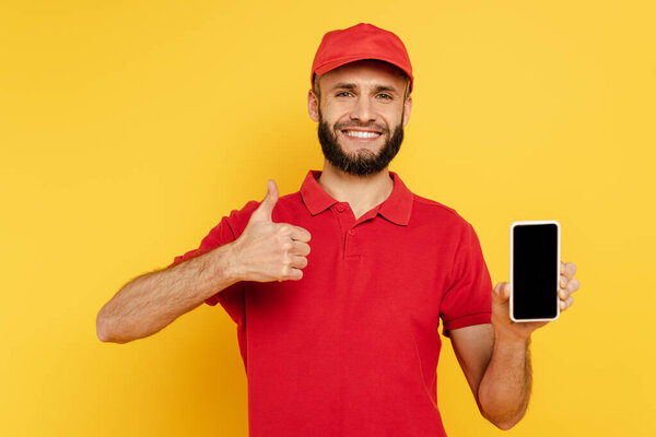 smiling bearded delivery man in red uniform with smartphone showing thumb up on yellow