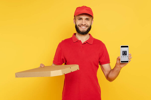 KYIV, UKRAINE - MARCH 30, 2020: smiling bearded delivery man in red uniform with pizza box showing smartphone with uber app on yellow