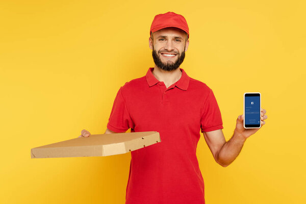 KYIV, UKRAINE - MARCH 30, 2020: smiling bearded delivery man in red uniform with pizza box showing smartphone with Facebook app on yellow