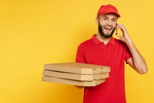 笑顔で髭を生やした配達人が赤い制服にピザボックスを着て黄色でスマホで話してる — ストック写真