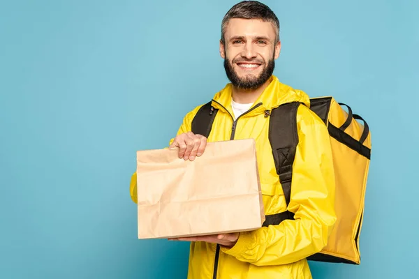 Usmívající Doručovatel Žluté Uniformě Batohem Drží Papírový Balíček Modrém Pozadí — Stock fotografie