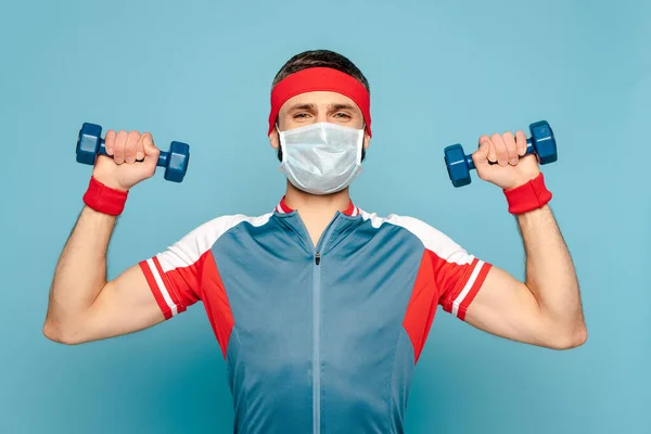 stylish sportsman in medical mask exercising with dumbbells on blue background