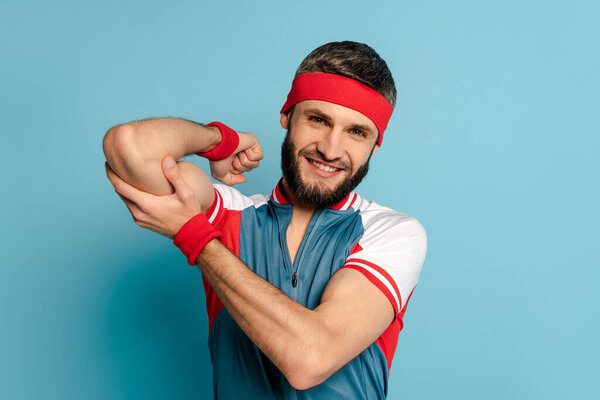 Улыбающийся стильный спортсмен трогает бицепсы на синем фоне
