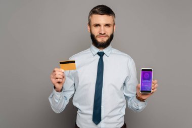 Yakışıklı sakallı iş adamı, elinde kredi kartı ve gri arka planda online alışveriş uygulaması olan akıllı telefon tutuyor.