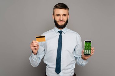 Yakışıklı sakallı iş adamı kredi kartı ve gri arka planda sağlık uygulamalı akıllı telefon tutuyor.