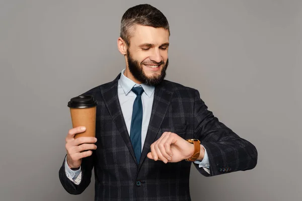 smiling handsome bearded businessman in suit with coffee to go looking at wristwatch on grey background