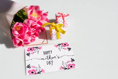 Küçük hediye kutularının yanında bir buket lale ve üzerinde mutlu anneler günü yazılı tebrik kartı.