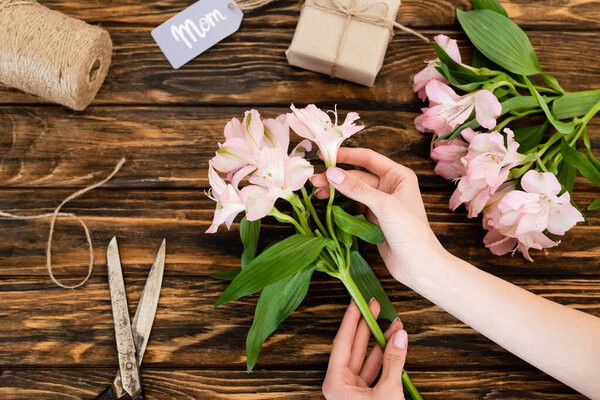 top view of woman holding pink flowers near scissors, jute twine rope and gift box with mom lettering on tag, mothers day concept 